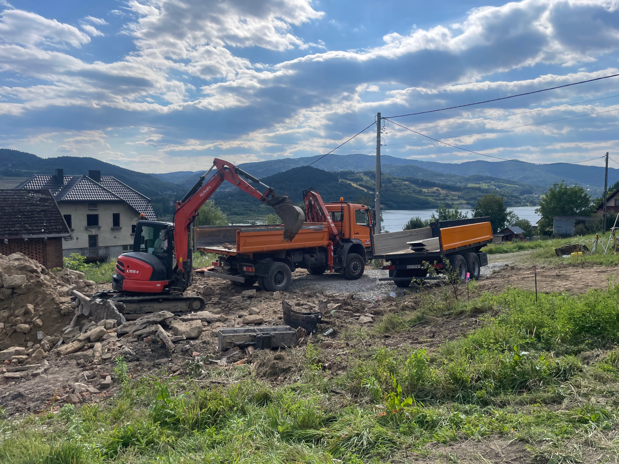 Czerwona koparka ładuje gruz do pomarańczowej ciężarówki na placu budowy z widokiem na góry i jezioro w tle, pod błękitnym niebem z obłokami.