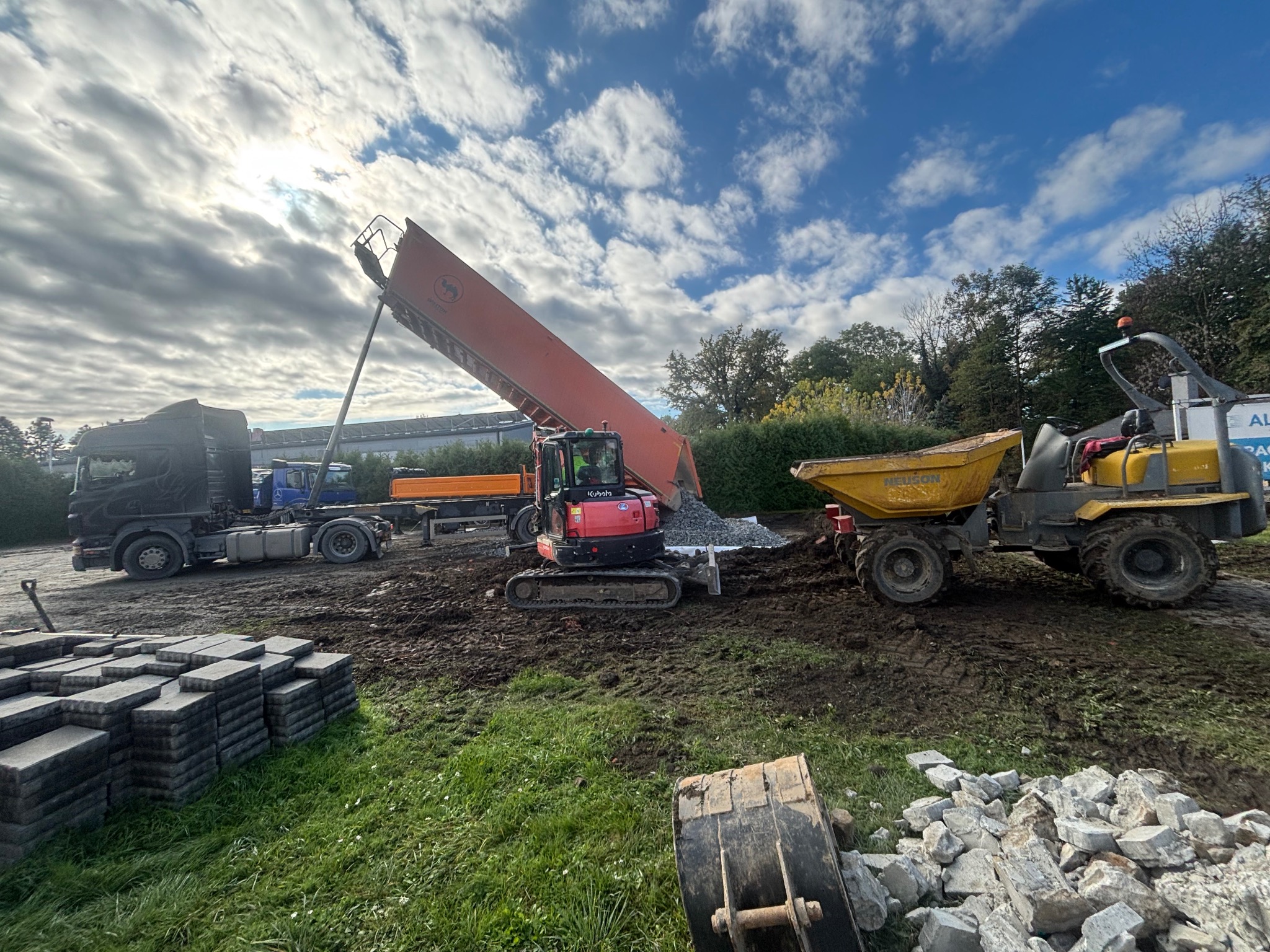 Ciężarówka wyładowuje gruz obok mini koparki Kubota i wywrotki Neuson. W tle zaparkowane pojazdy i ułożona kostka brukowa. Prace ziemne w toku.