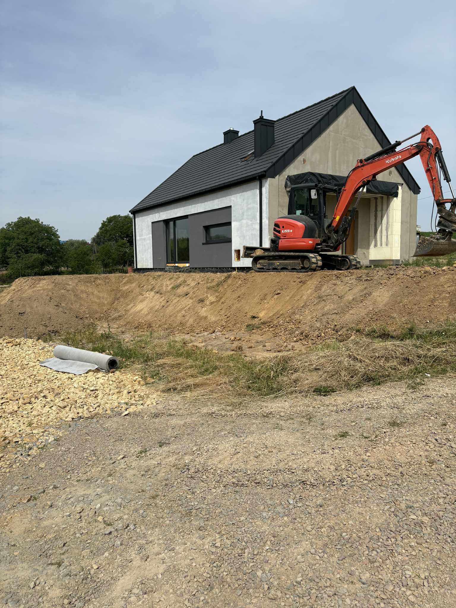 Czerwona mini koparka Kubota na wzgórzu ziemi przed nowoczesnym domem w trakcie budowy. W tle drzewa i niebo. Na pierwszym planie rolka geowłókniny.