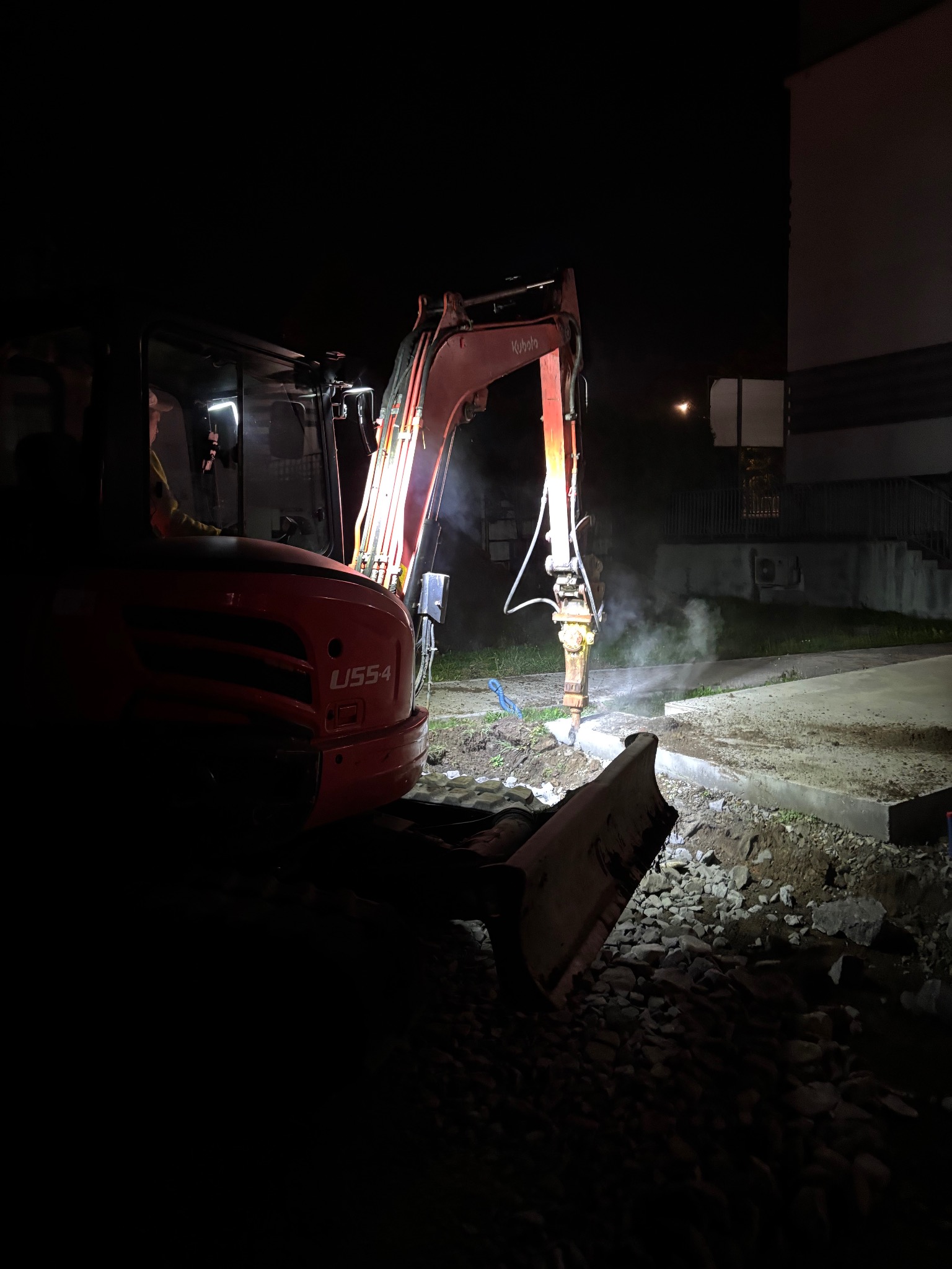 Czerwona koparka Kubota U55-4 z włączonymi światłami pracuje nocą, rozbijając betonowy fundament. Widoczne gruz i pył. Praca w toku.