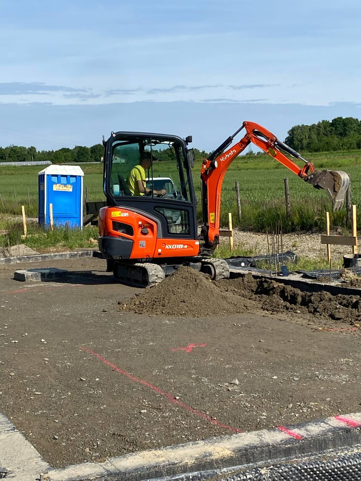 Pomarańczowa mini koparka Kubota KX018-4 w trakcie prac ziemnych na budowie, z widocznym operatorem i przenośną toaletą w tle.