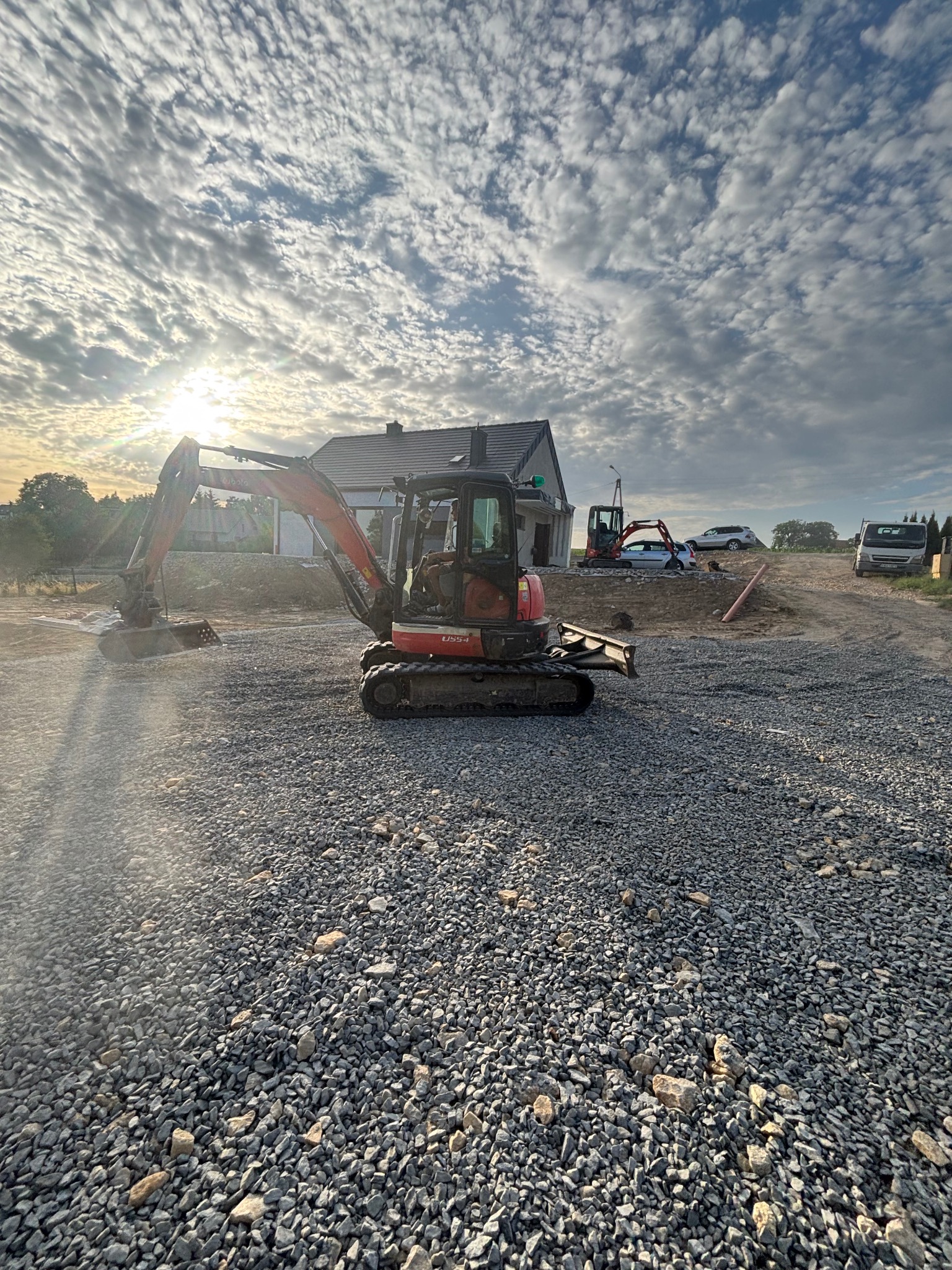 Czerwony minikoparka na żwirowym podłożu, w tle dom w budowie i błękitne niebo z chmurami. Maszyny budowlane w oddali. Słoneczny dzień.