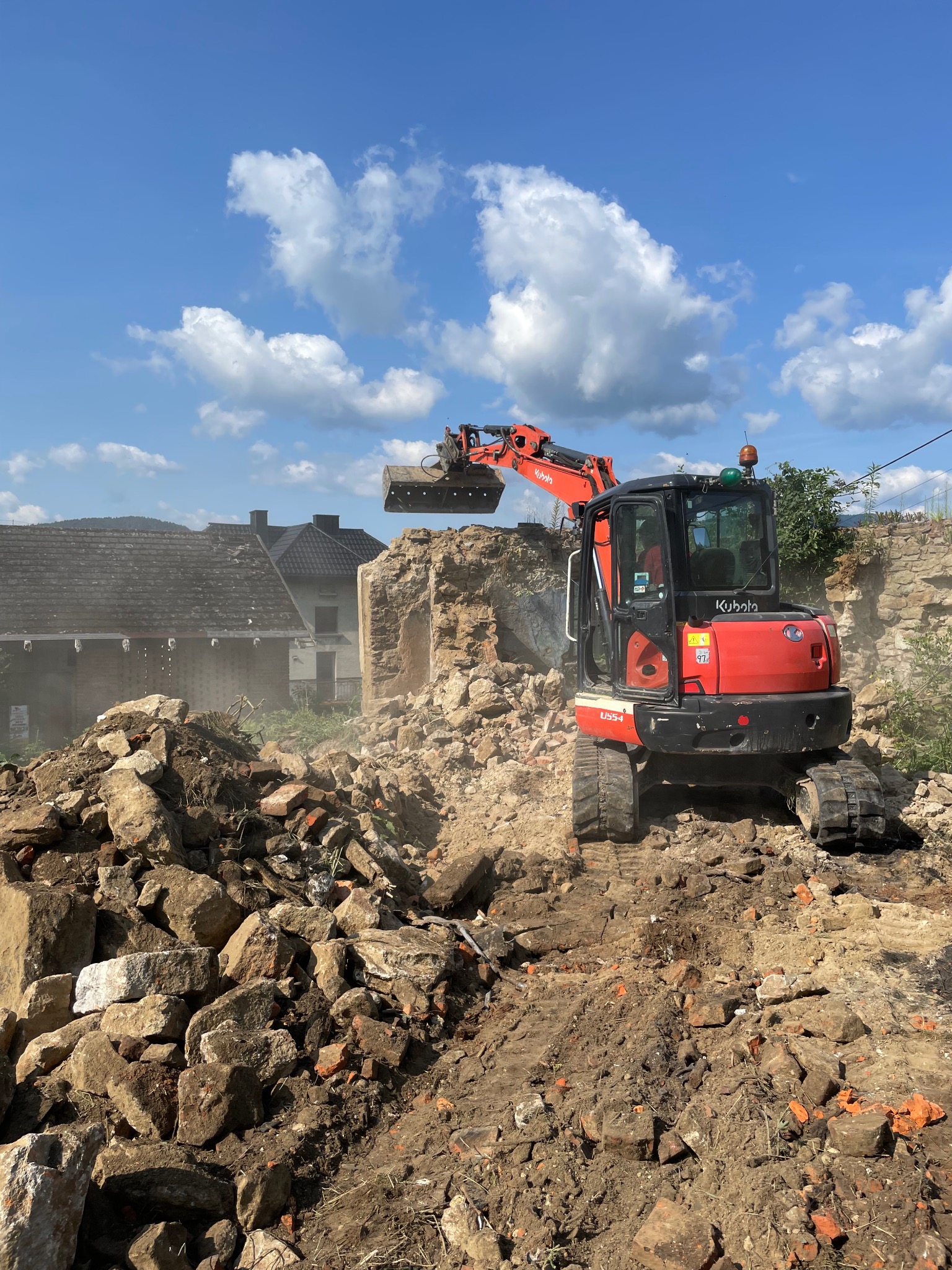 Czerwona mini koparka Kubota U55-4 w trakcie rozbiórki kamiennego muru na tle błękitnego nieba z chmurami. Gruz i ziemia na pierwszym planie.