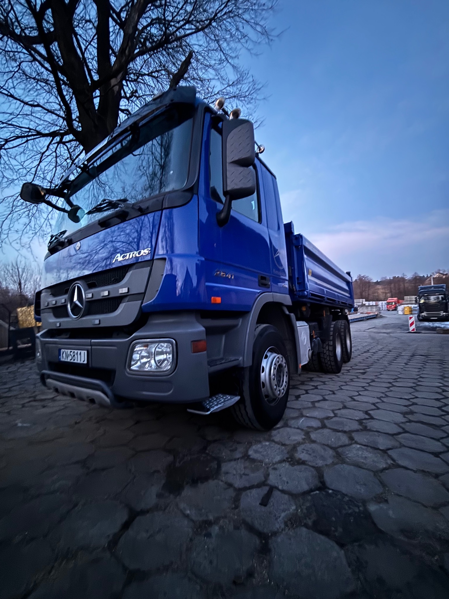 Niebieski wywrotka Mercedes Actros 2641 na brukowanym placu, widok z dołu, z drzewem w tle i elementami placu budowy w oddali.