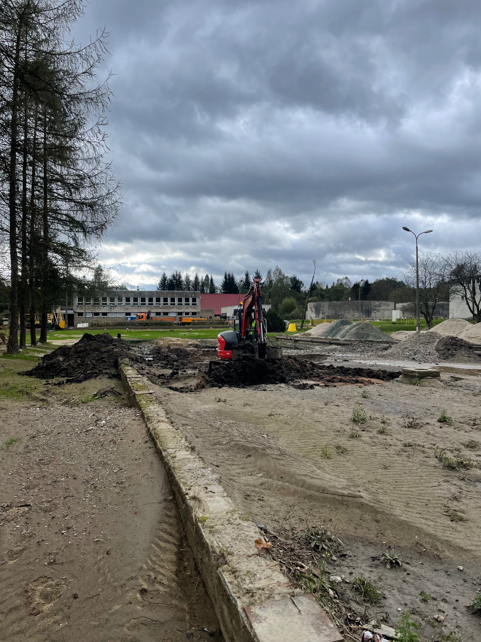 Czerwony minikoparka w trakcie prac ziemnych na placu budowy, widoczne hałdy ziemi i gruzu, w tle budynek i pochmurne niebo. Prace przygotowawcze pod inwestycję.