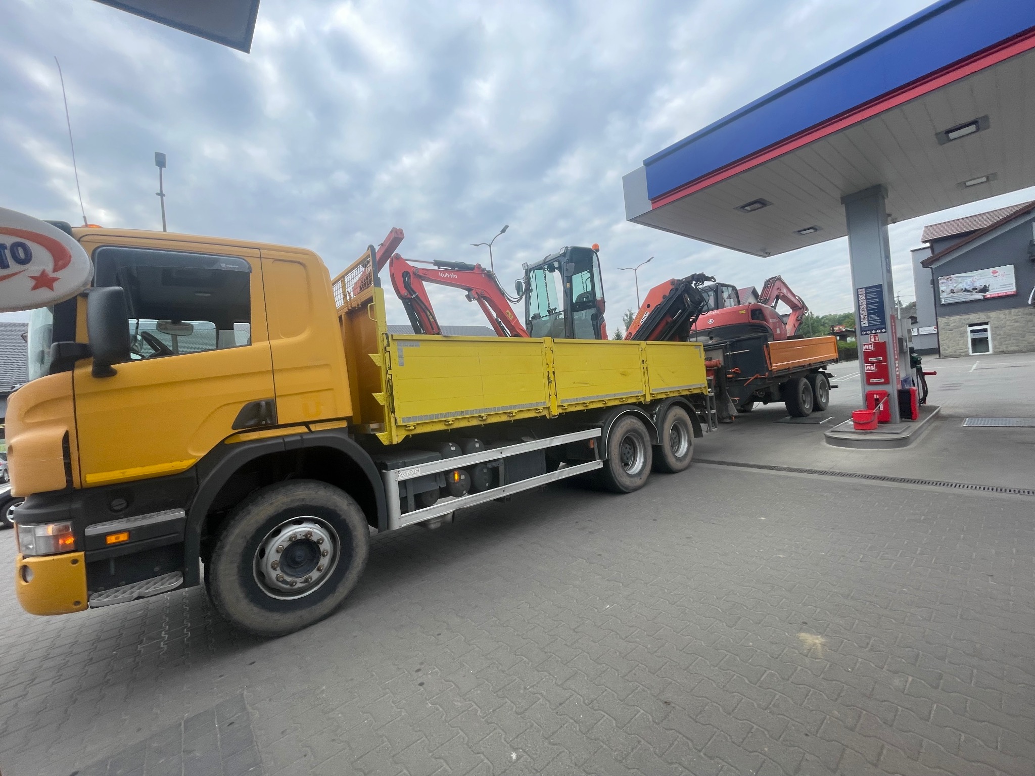Żółta ciężarówka z dwoma małymi koparkami na pace, stojąca na stacji benzynowej. Widoczne detale: opony ciężarówki, czerwone koparki, dystrybutor paliwa.