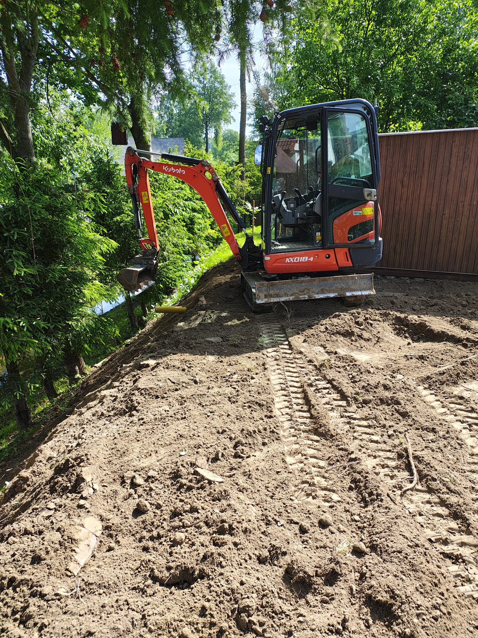 Pomarańczowa mini koparka Kubota KX018-4 na nierównym terenie z ziemią, widoczne ślady gąsienic, zieleń w tle, brązowe ogrodzenie po prawej.