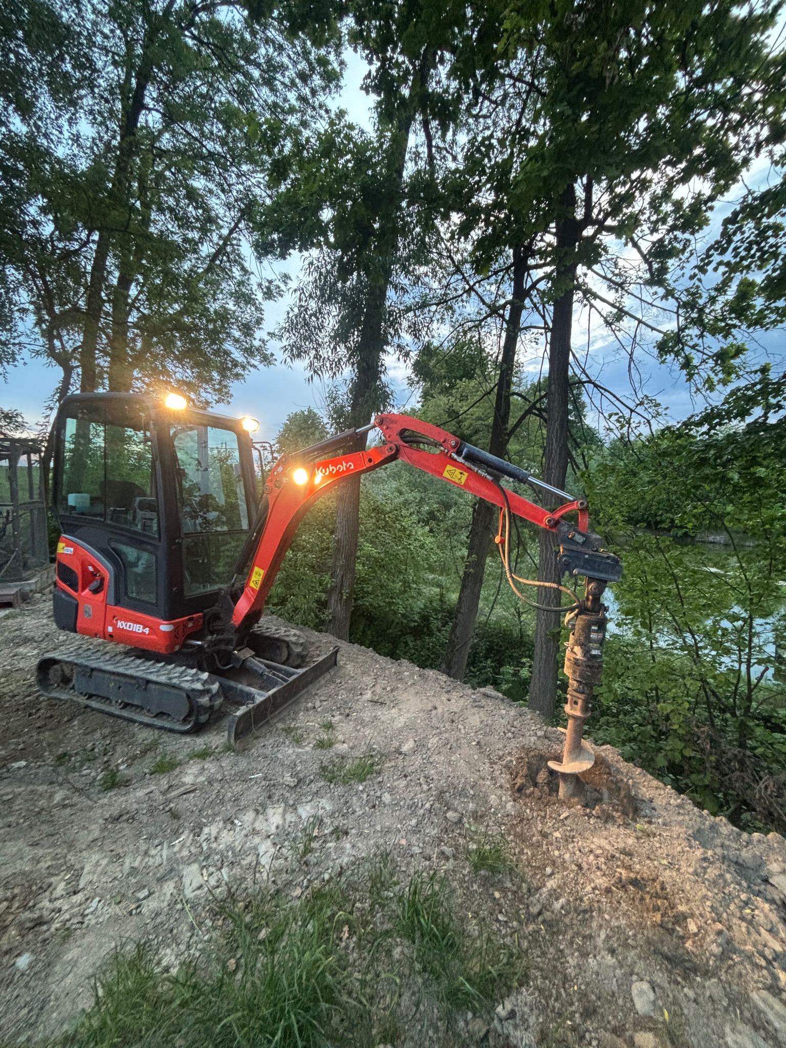 Czerwona mini koparka Kubota z wiertłem świdrowym w trakcie pracy na wzgórzu, otoczona drzewami. Widoczne oświetlenie robocze i fragment jeziora w tle.