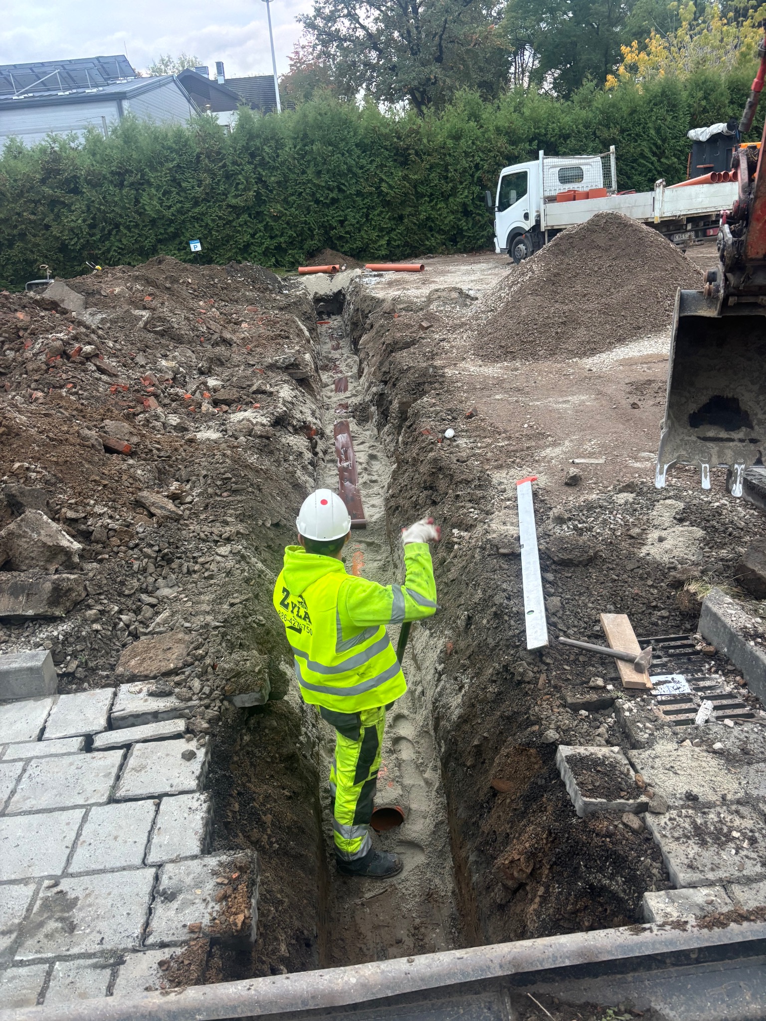 Pracownik w żółtym ubraniu roboczym stoi w wykopie z rurami. W tle widać koparkę, ciężarówkę i hałdę ziemi. Prace ziemne w toku, widok zza pleców.