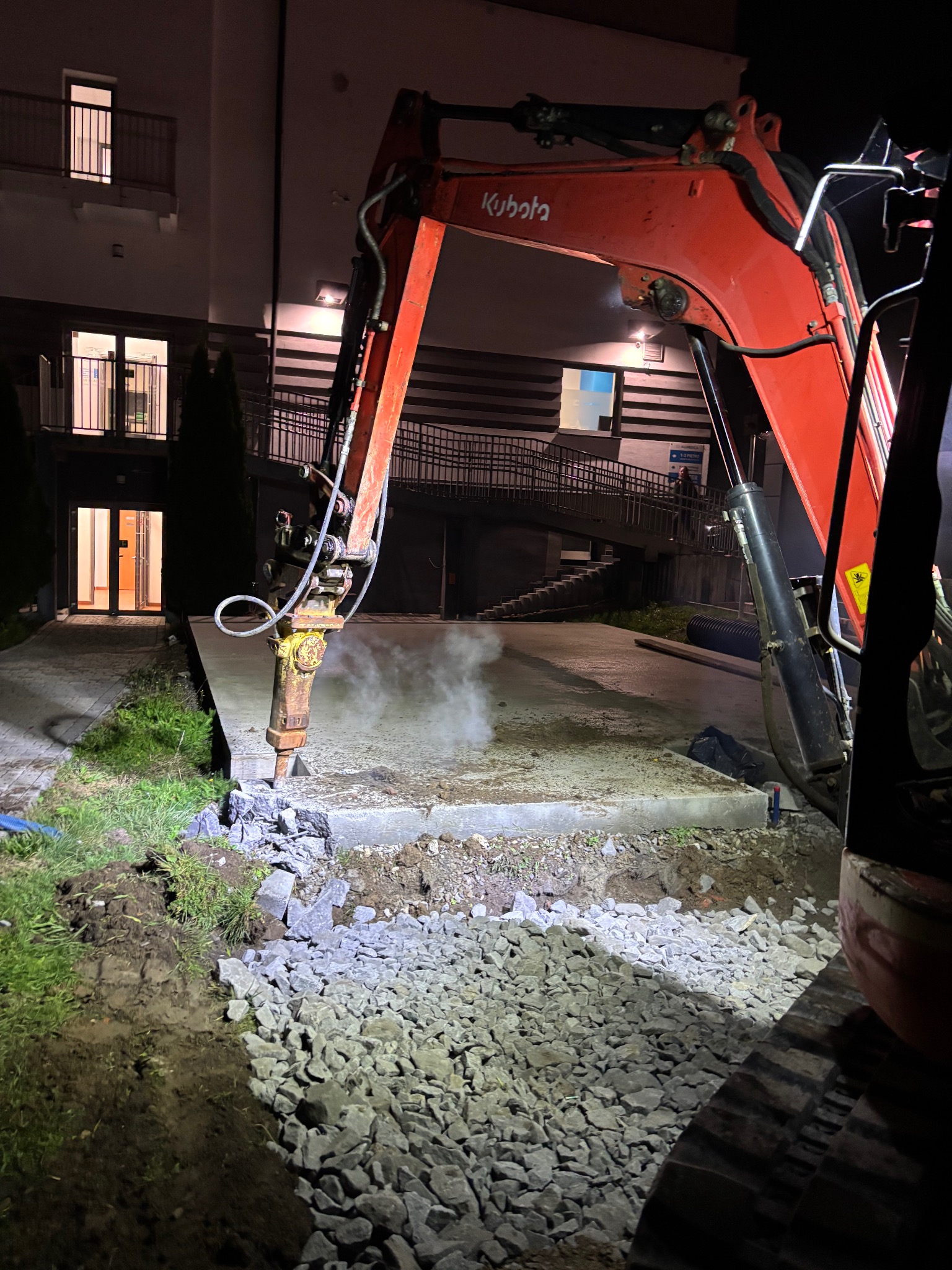 Czerwony minikoparka Kubota z młotem pneumatycznym kruszy betonową płytę na zewnątrz budynku mieszkalnego, przygotowując teren pod dalsze prace.