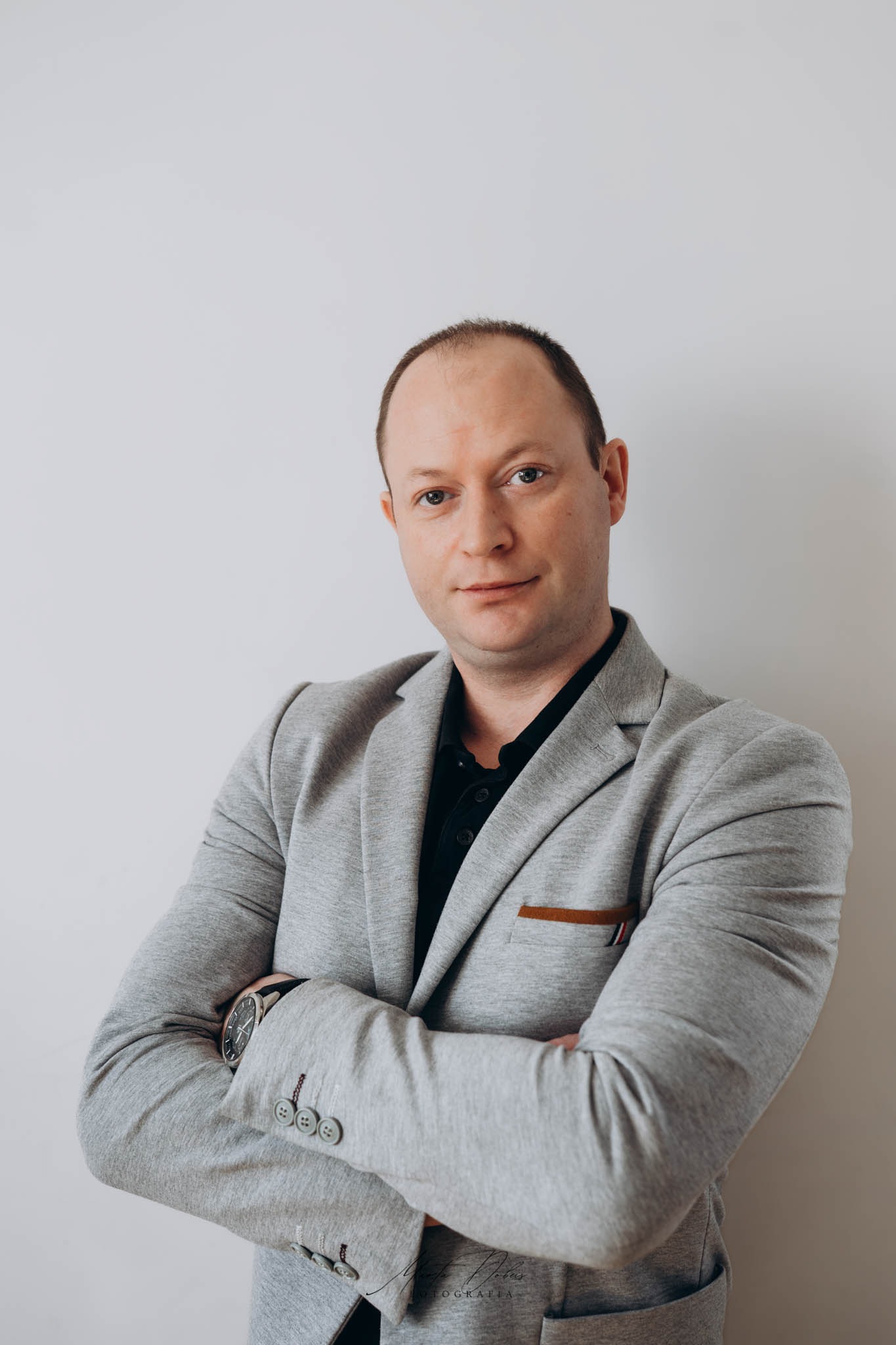 Portret mężczyzny w szarym blezerze, z zegarkiem na ręku, na jasnym tle. Uśmiechnięty, z założonymi rękami. Profesjonalny wizerunek.