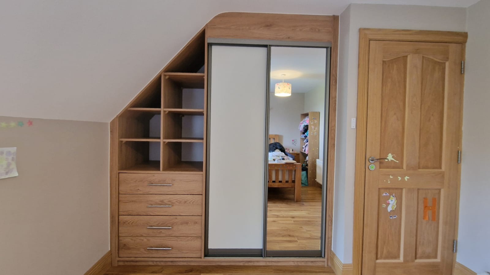 Zabudowa garderoby z drewna pod skosem, z przesuwnymi drzwiami lustrzanymi, obok drewniane drzwi z naklejkami w pokoju dziecięcym.