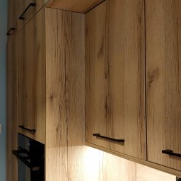 IMPART Adam Malina - Drewniane szafki kuchenne z czarnymi uchwytami i oświetleniem LED, z piekarnikiem w zabudowie. Fronty z wyraźnym rysunkiem słojów, ciepłe światło podkreśla fakturę drewna.