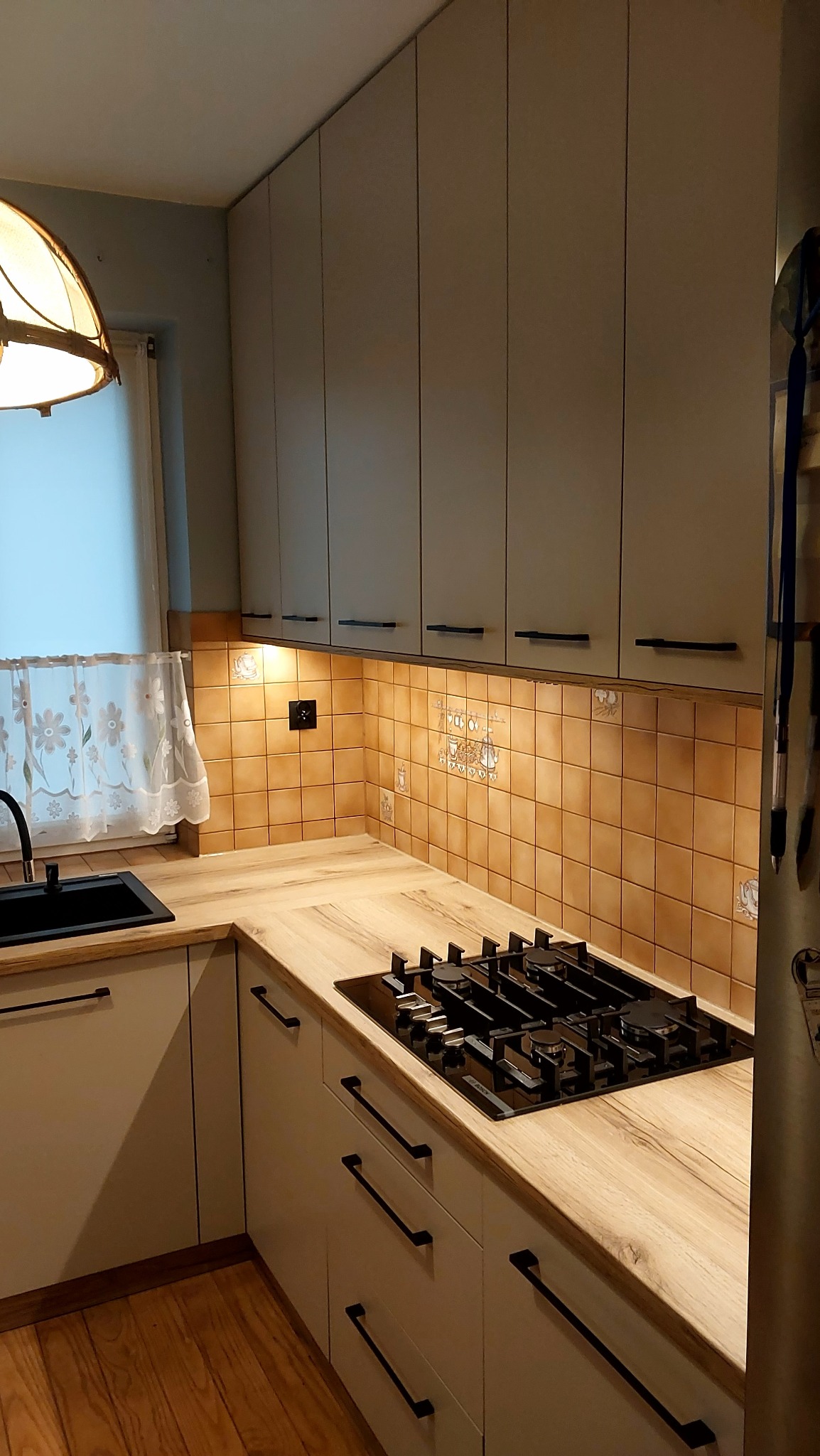 Kremowa kuchnia z drewnianym blatem i brązowymi płytkami. Uwagę przykuwa czarna płyta gazowa i długie, czarne uchwyty szafek. Nad zlewem wisi krótka firanka.