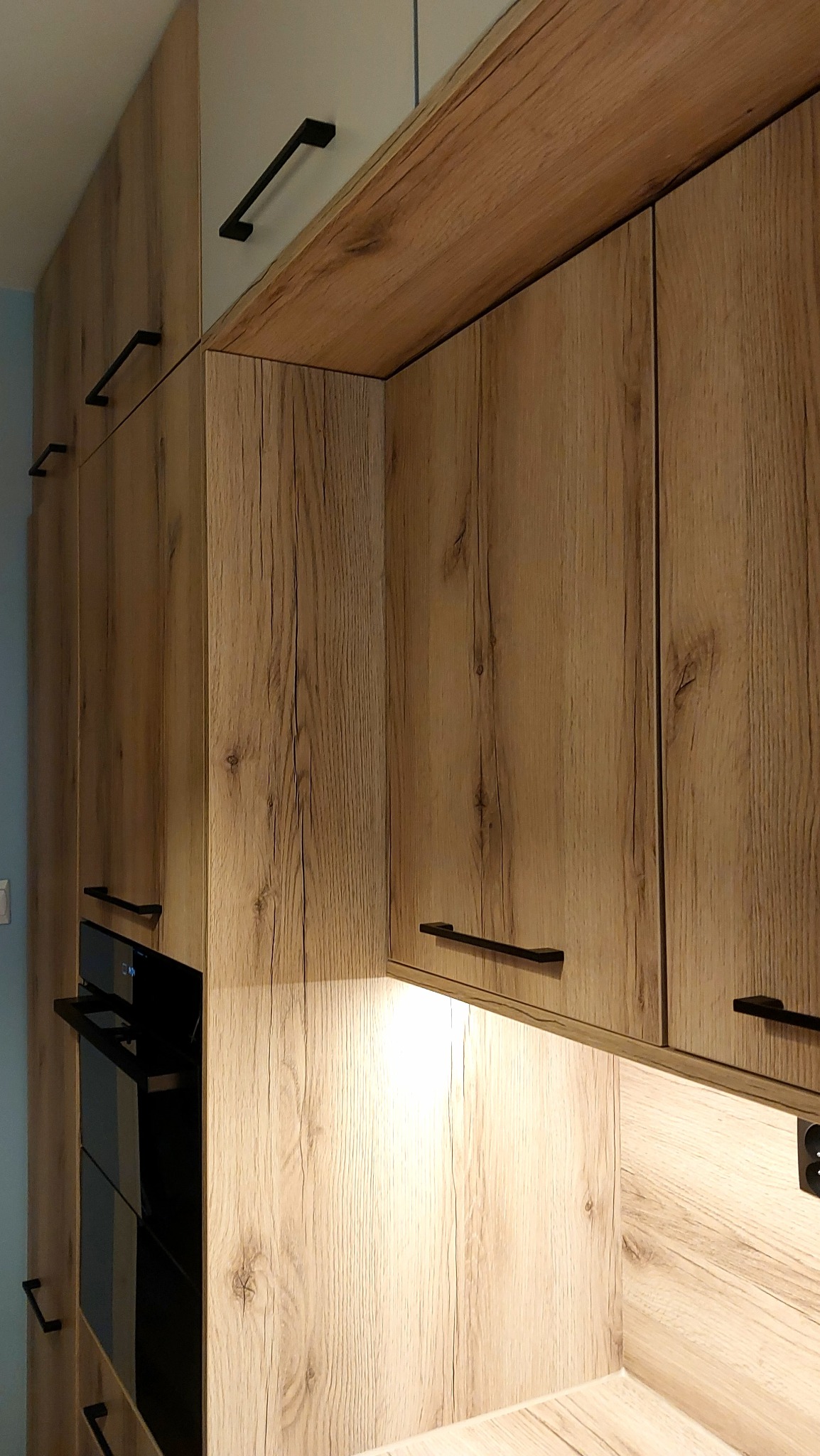 Drewniane szafki kuchenne z czarnymi uchwytami i oświetleniem LED, z piekarnikiem w zabudowie. Fronty z wyraźnym rysunkiem słojów, ciepłe światło podkreśla fakturę drewna.