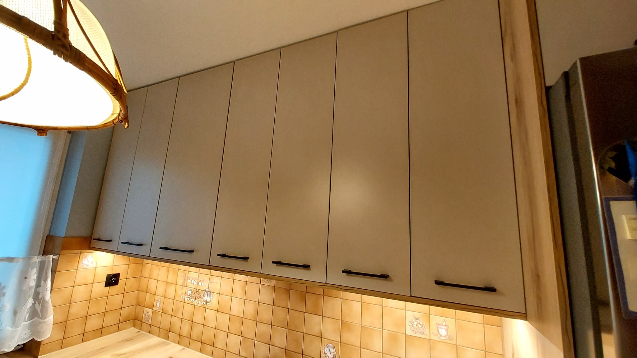 Beżowe szafki kuchenne wiszące z czarnymi uchwytami, oświetlone od dołu, z widoczną mozaiką na ścianie i fragmentem lampy w rogu kadru.