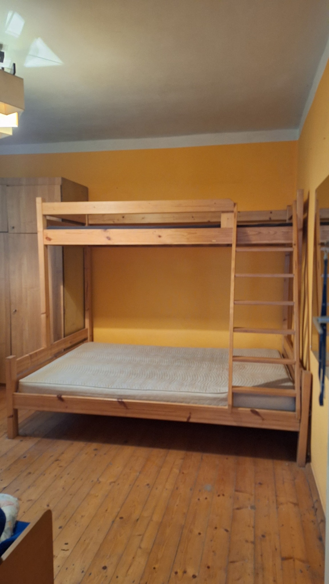 Drewniane łóżko piętrowe z drabinką w pokoju z żółtymi ścianami i drewnianą podłogą. Obok szafa z drewna. Widok na całą konstrukcję łóżka.