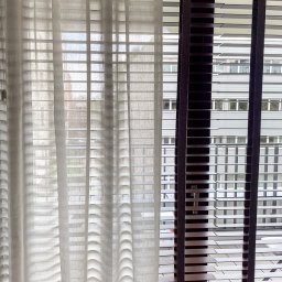 Pasadena home & deco - Jasna, delikatna firana z efektem falowania obok ciemnych żaluzji drewnianych z widokiem na budynek za oknem. Ujęcie z bliska, z logo firmy w lewym dolnym rogu.