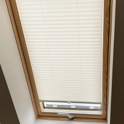 Pasadena home & deco - Jasna plisa okienna zamontowana w drewnianej ramie okna dachowego, widoczny mechanizm sterujący. Neutralne tło, stonowana kolorystyka.