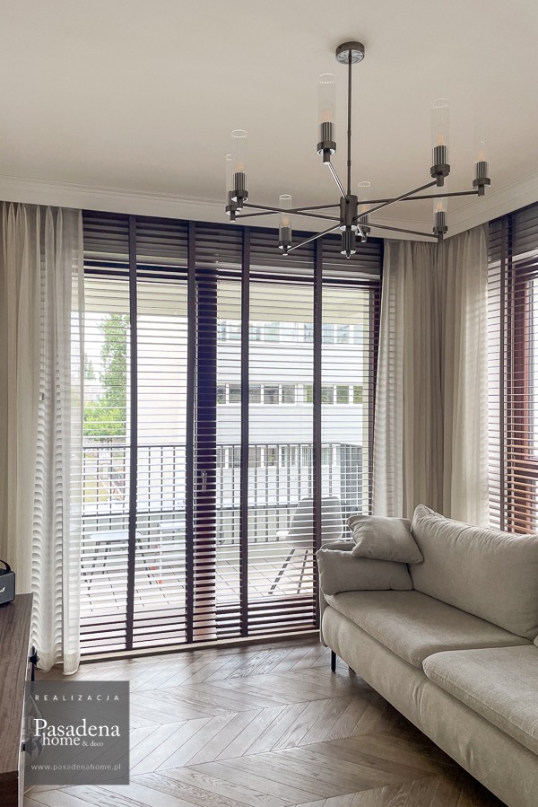 Elegancki salon z drewnianymi żaluzjami w kolorze Tiger Eye, zwiewnymi firanami i nowoczesnym żyrandolem. Parkiet w jodełkę dodaje wnętrzu klasy.