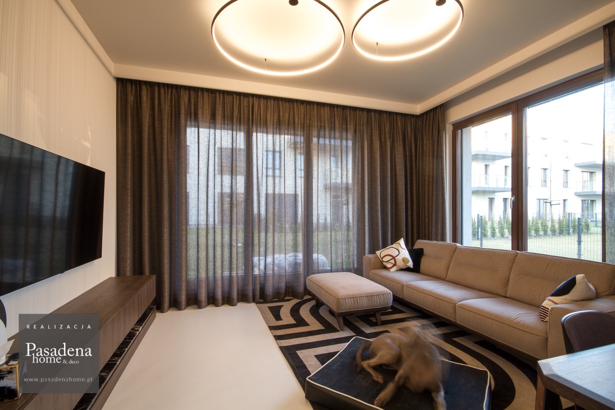 Nowoczesny salon z miedzianymi zasłonami o splocie plecionki, szyny sufitowe KS białe. Widoczny fragment sofy, stolika i dywanu w geometryczne wzory.