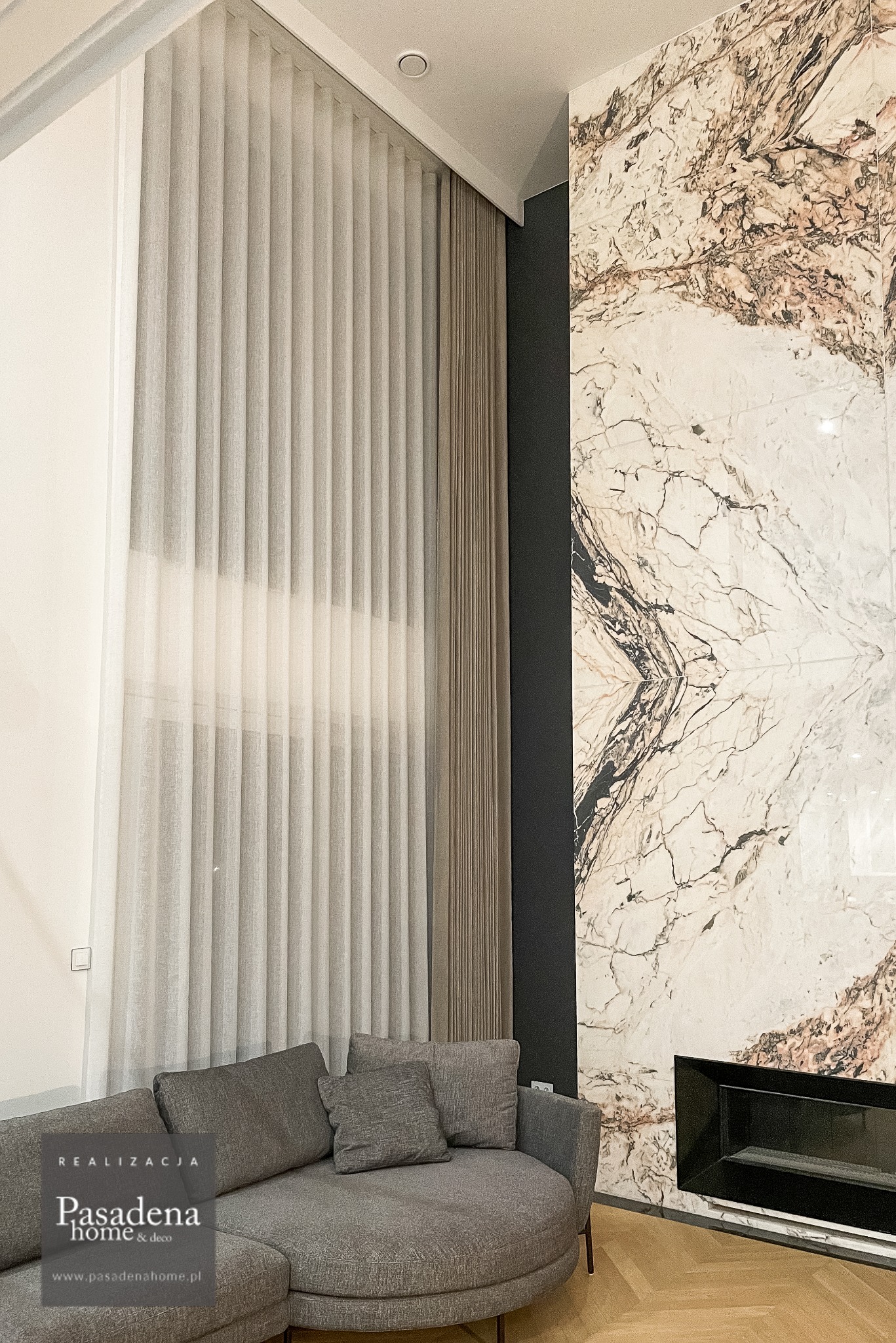 Wysokie okno z delikatną, półprzezroczystą firaną i zasłoną w ciepłym odcieniu piasku, tworzące elegancką dekorację okienną na dwóch kondygnacjach. Marmurowa ściana obok.