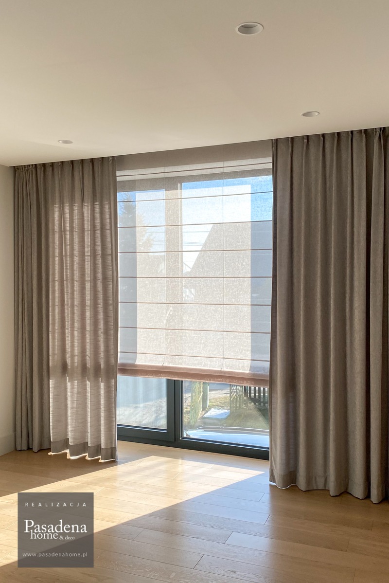 Jasne wnętrze z roletą rzymską i zasłonami w odcieniach szarości. Delikatna faktura materiałów, naturalne światło podkreśla elegancję aranżacji okiennej.