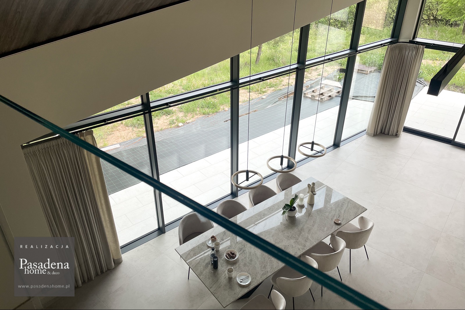 Wysokie okna z beżowymi zasłonami Broadway 17 na czarnych karniszach Loft 25, widok z góry na jadalnię z marmurowym stołem i krzesłami. Lekki i elegancki wystrój.