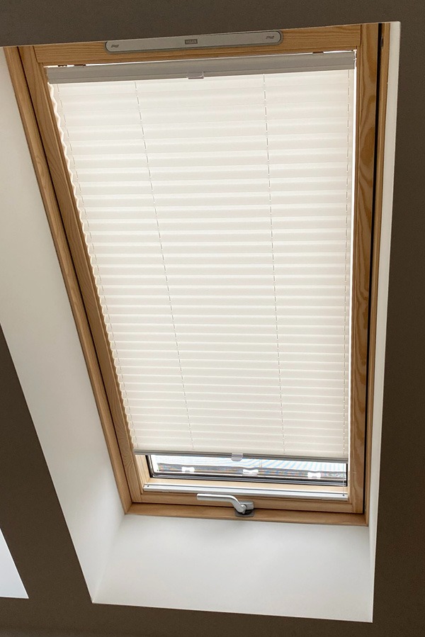 Jasna plisa okienna zamontowana w drewnianej ramie okna dachowego, widoczny mechanizm sterujący. Neutralne tło, stonowana kolorystyka.