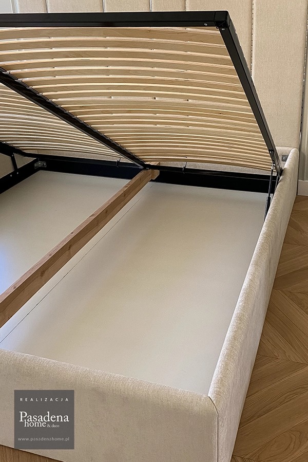 Tapicerowane łóżko z podnoszonym stelażem i pojemnikiem na pościel, widok z góry. Beżowa tkanina, drewniany stelaż, parkiet na podłodze. Widoczny fragment tapicerowanej ściany.