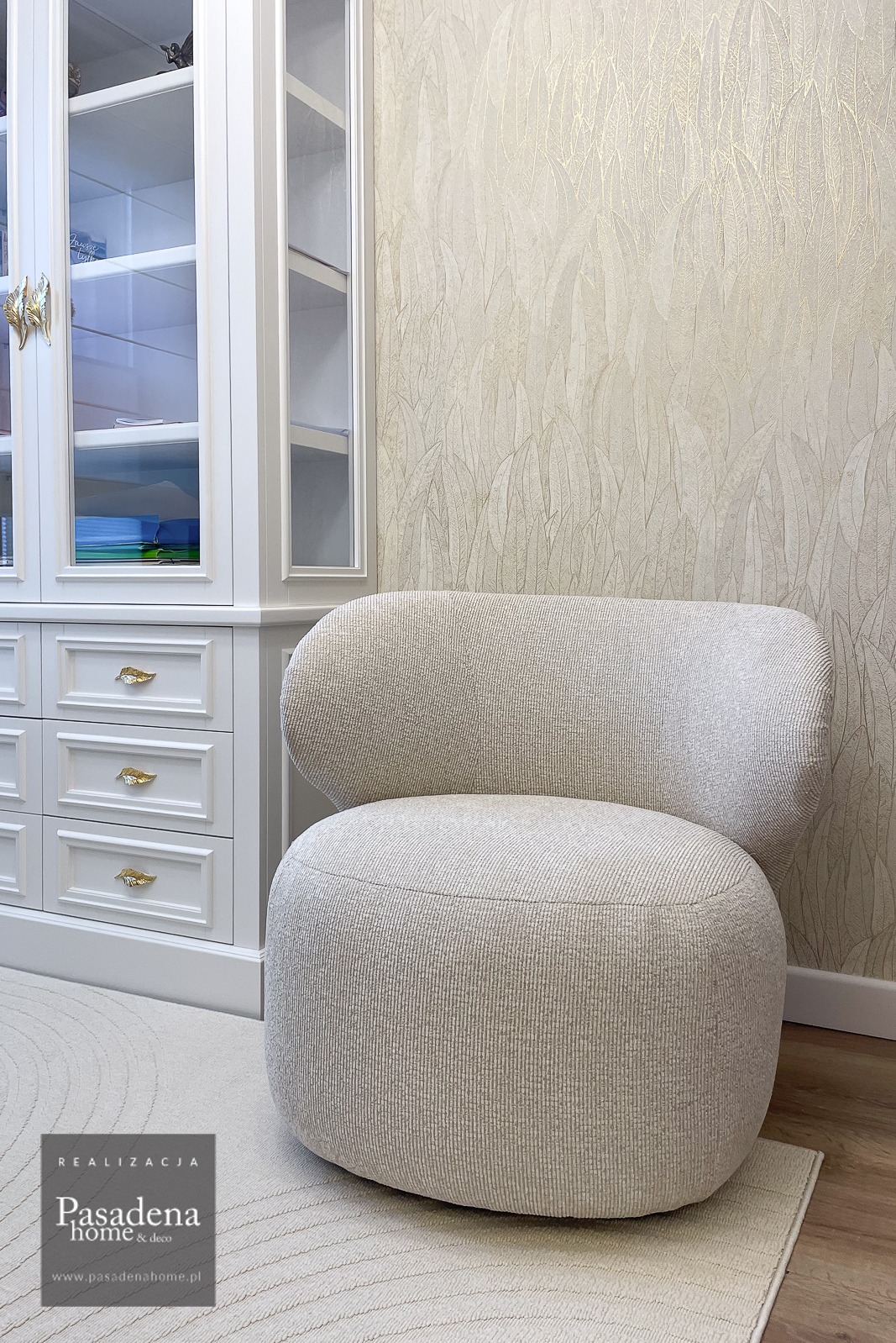 Beżowy fotel Riley z tapicerką w prążki, na tle ściany w liście i białej komody z przeszkloną witryną. Widoczne logo firmy Pasadena Home & deco.