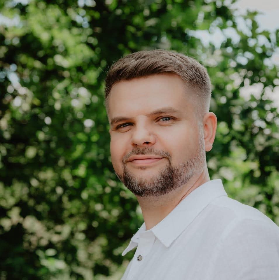 Portret uśmiechniętego mężczyzny z brodą, w białej koszuli, na tle zieleni. Ujęcie z bliska, z delikatnym rozmyciem tła.