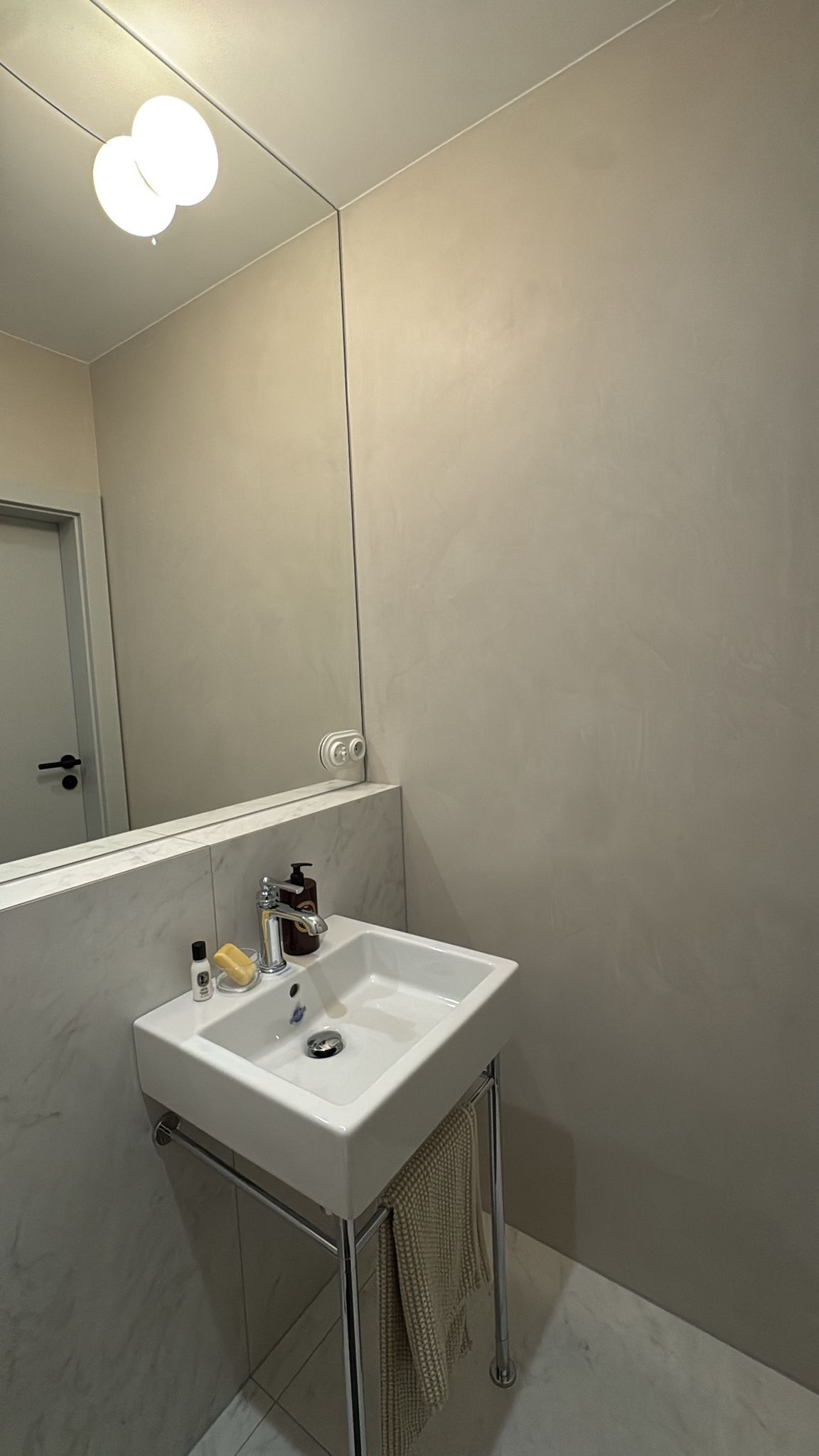 Minimalistyczna łazienka z umywalką na metalowym stelażu, lustrem bez ramy i ścianą wykończoną w odcieniach beżu. Widoczne akcesoria łazienkowe i ręcznik.