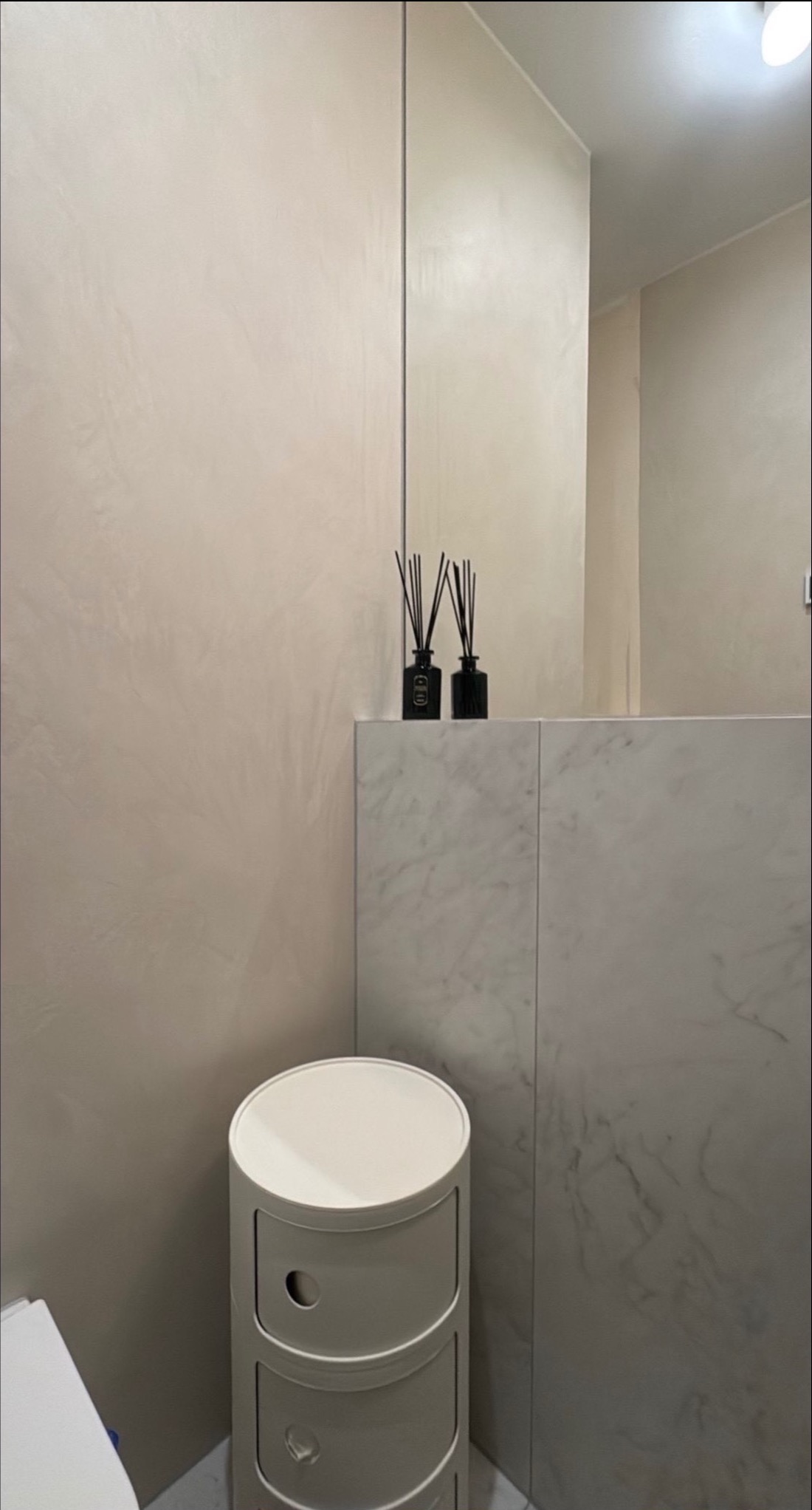 Beżowa ściana z efektem dekoracyjnym w łazience, biała szafka z szufladami i marmurowy blat z czarnymi dyfuzorami zapachowymi. Minimalistyczny design.