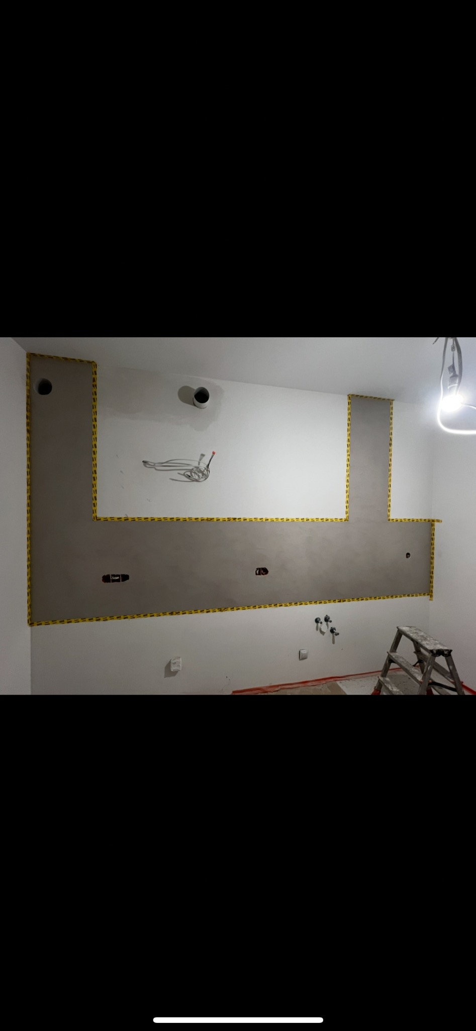 Ściana w trakcie szpachlowania z wyznaczonym obszarem, zabezpieczona żółtą taśmą. Widoczne instalacje, drabina i surowy stan wykończenia wnętrza.