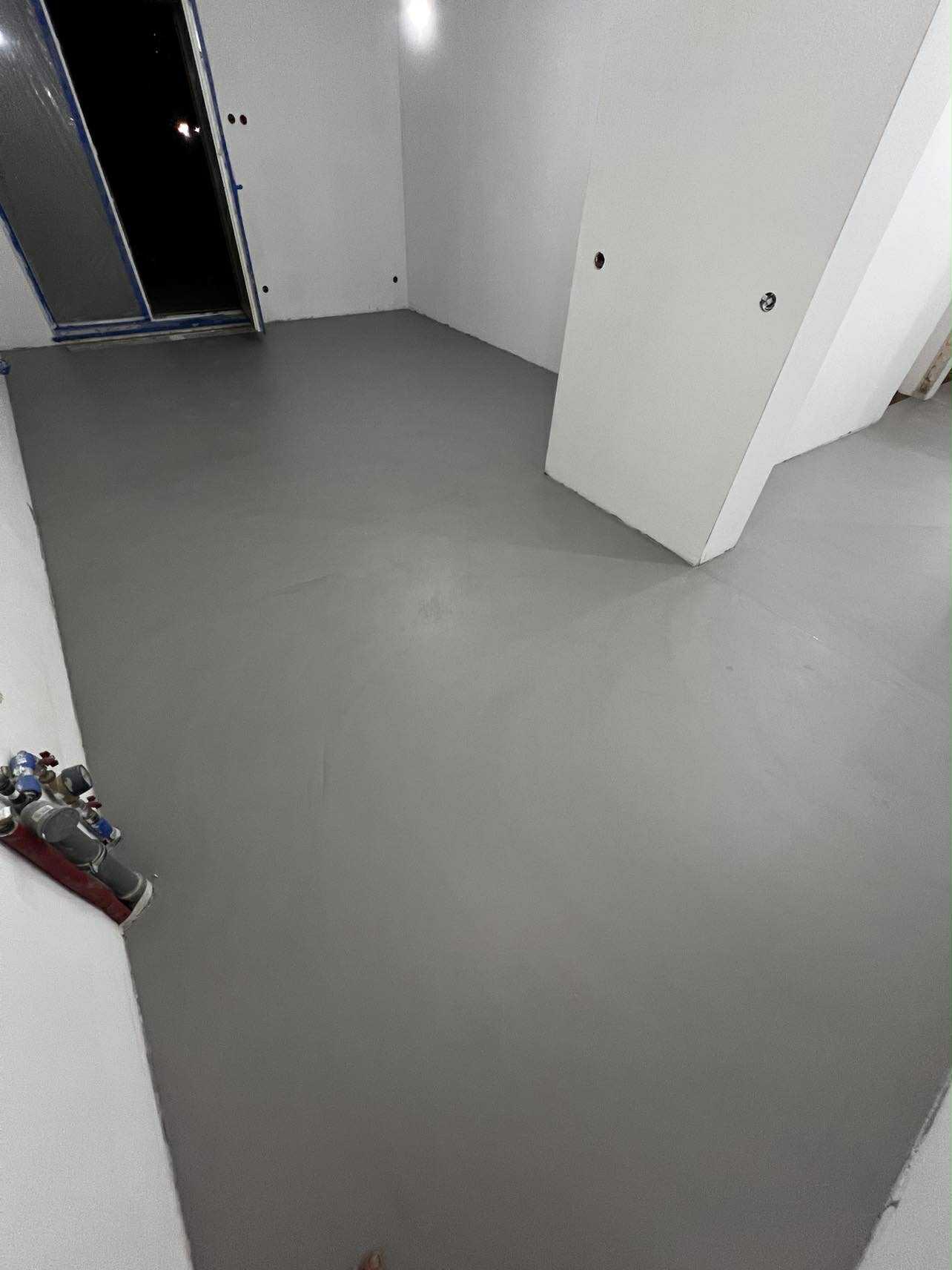 Szara, gładka posadzka w surowym wnętrzu z widocznymi instalacjami przy ścianie. Minimalistyczny design, świeżo wylana powierzchnia, gotowa do dalszych prac wykończeniowych.
