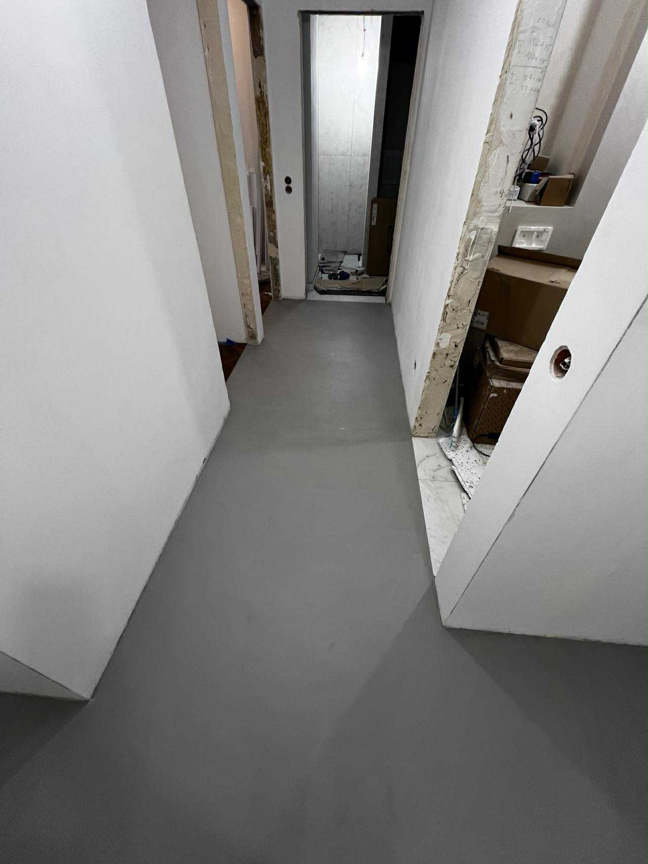 Szara, gładka posadzka w korytarzu w trakcie remontu. Widoczne surowe ściany i przygotowania do dalszych prac wykończeniowych. Minimalistyczny design.