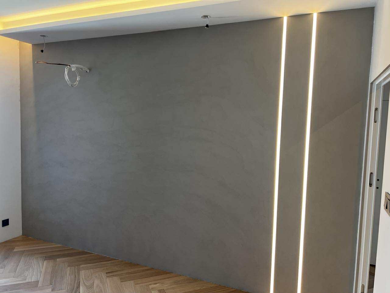 Ściana z efektem betonu architektonicznego, oświetlona pionowymi pasami LED, parkiet w jodełkę. Minimalistyczny design wnętrza z widocznymi instalacjami.