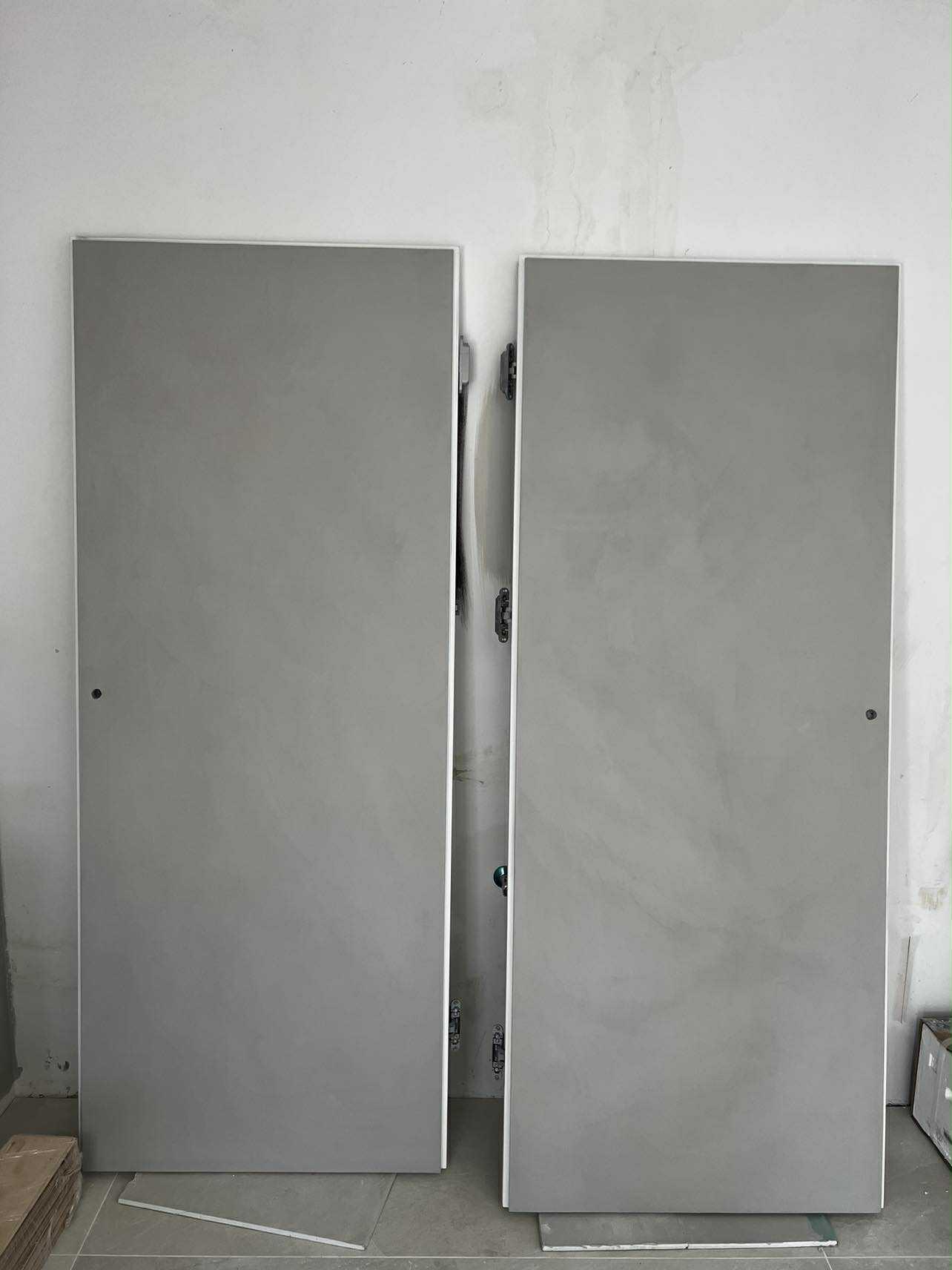 Dwa szare, matowe panele drzwiowe o gładkiej fakturze, oparte o ścianę w trakcie montażu. Widoczne zawiasy i surowy stan wykończenia wnętrza.