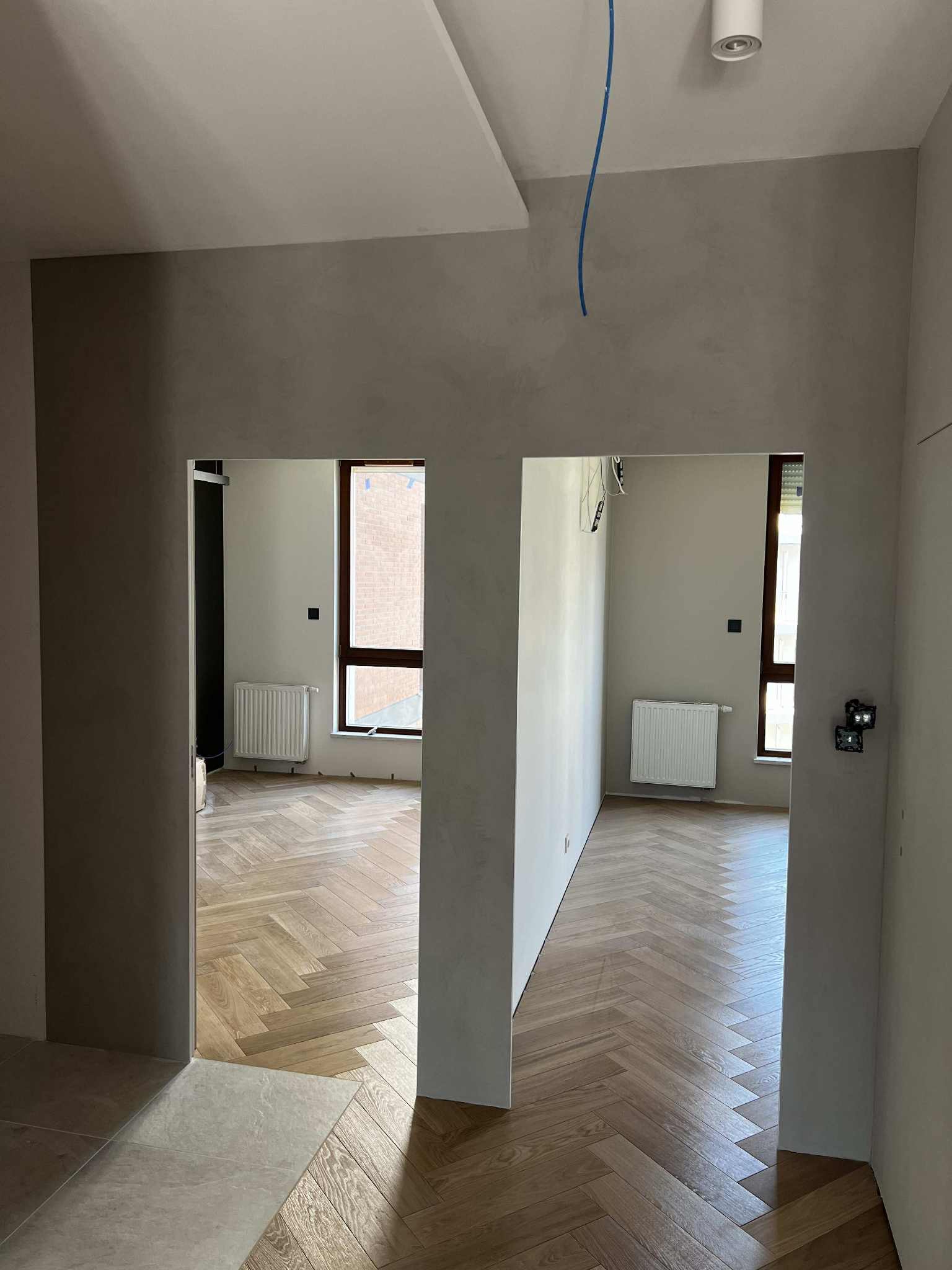 Jasne parkiety ułożone w jodełkę w nowym mieszkaniu, widoczne przez dwa przejścia. Ściany w odcieniach szarości, grzejniki pod oknami. Niebieski kabel zwisa z sufitu.