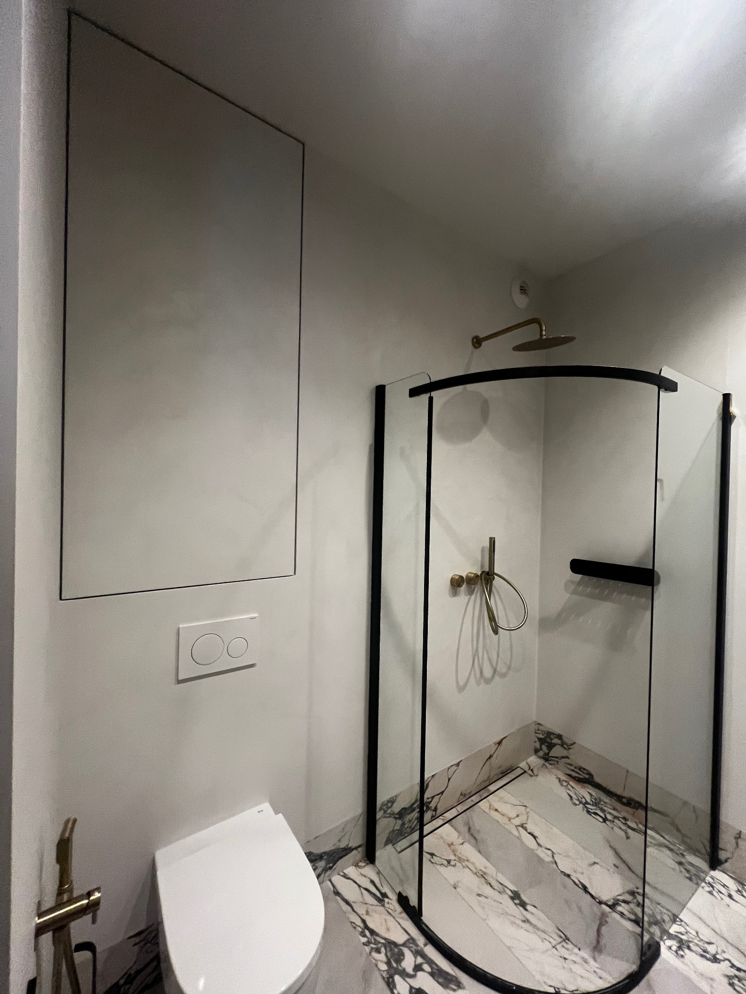 Elegancka łazienka z kabiną prysznicową typu walk-in, marmurową posadzką i ukrytą spłuczką. Armatura w kolorze złotym dodaje luksusowego charakteru.