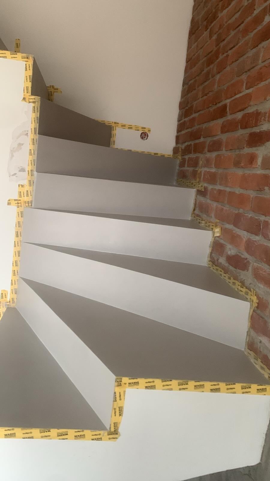 Schody po malowaniu w Kobyłce, zabezpieczone taśmą malarską, z fragmentem ściany z czerwonej cegły. Widok z góry, ukazujący detale wykończenia.