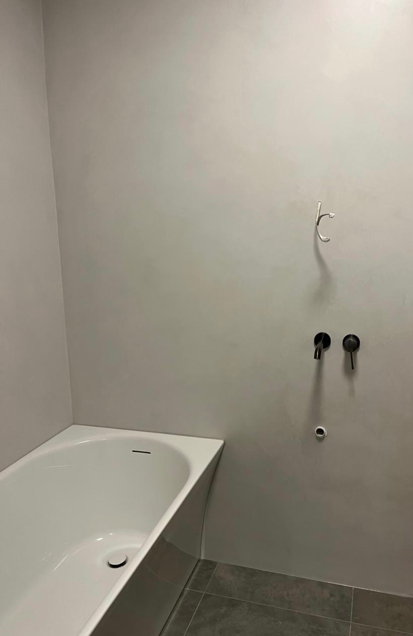 Minimalistyczna łazienka z wanną narożną i szarą ścianą wykończoną mikrocementem. Widoczne czarne baterie i płytki podłogowe w odcieniach szarości.