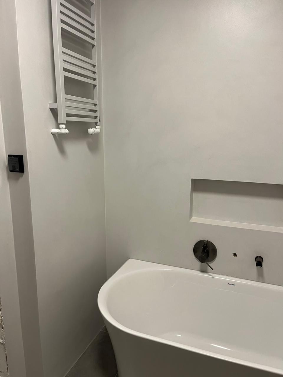 Minimalistyczna łazienka z białą wanną, grzejnikiem drabinkowym i szarą ścianą z wnęką. Armatura w kolorze ciemnego chromu. Nowoczesny design.