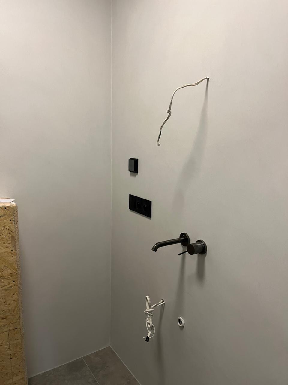 Szara ściana w łazience z czarną armaturą, gniazdkami i wystającymi kablami. Widoczny fragment podłogi z szarych płytek i płyta OSB w rogu.