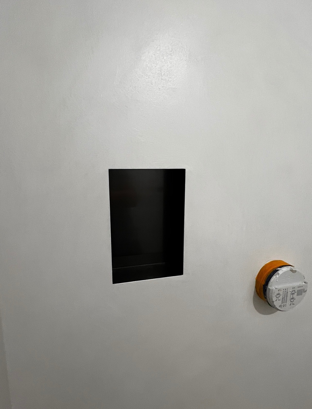 Biała ściana z wnęką wykończoną na czarno. Obok widoczny element instalacji zabezpieczony pomarańczową taśmą. Minimalistyczne wykończenie wnętrza.