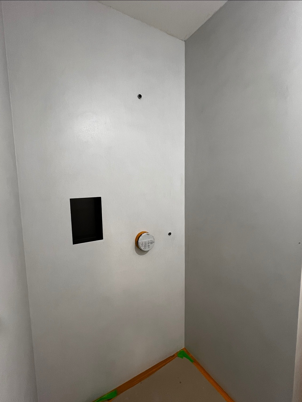 Gładkie, szare ściany w rogu pomieszczenia z otworem i puszką elektryczną. Podłoga zabezpieczona taśmą malarską. Minimalistyczne wykończenie wnętrza.