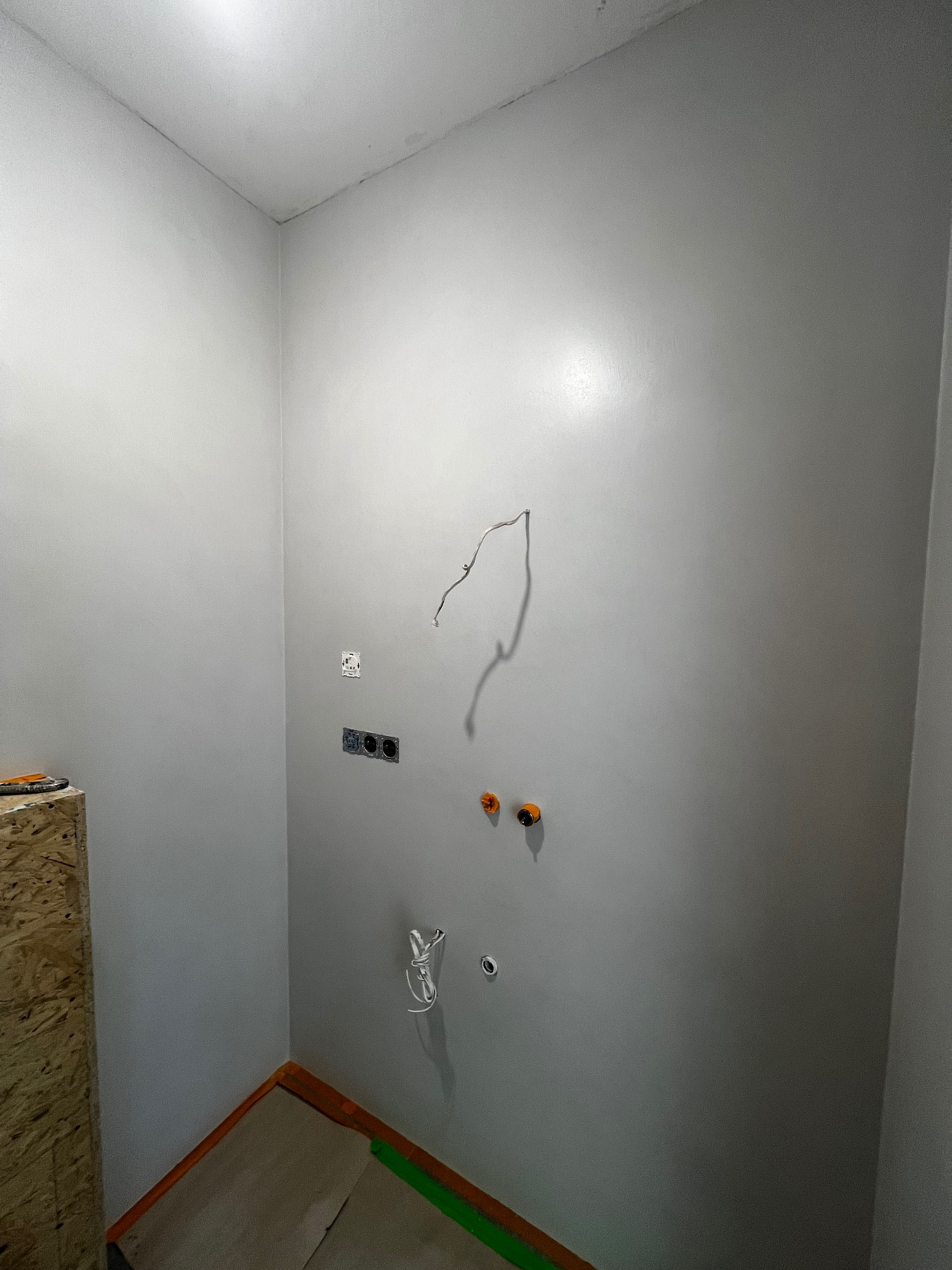 Świeżo pomalowane, gładkie ściany w rogu pomieszczenia z wystającymi przewodami elektrycznymi i instalacją wodną. Widoczne puszki elektryczne i fragment podłogi z zabezpieczeniem.