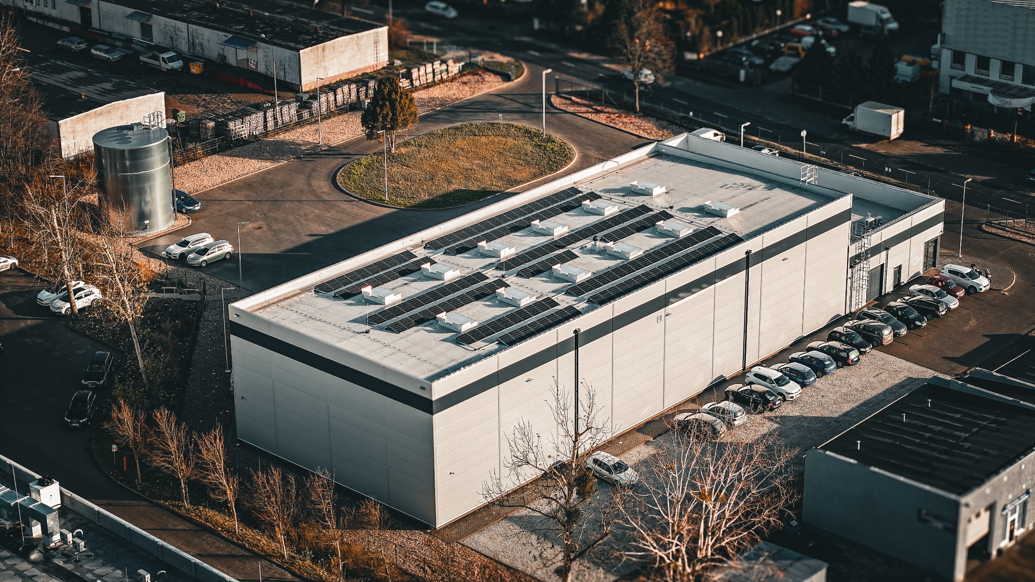 Widok z góry na duży budynek z panelami słonecznymi na dachu, otoczony parkingiem i zielenią. Ujęcie z efektem miniatury, podkreślające detale instalacji.