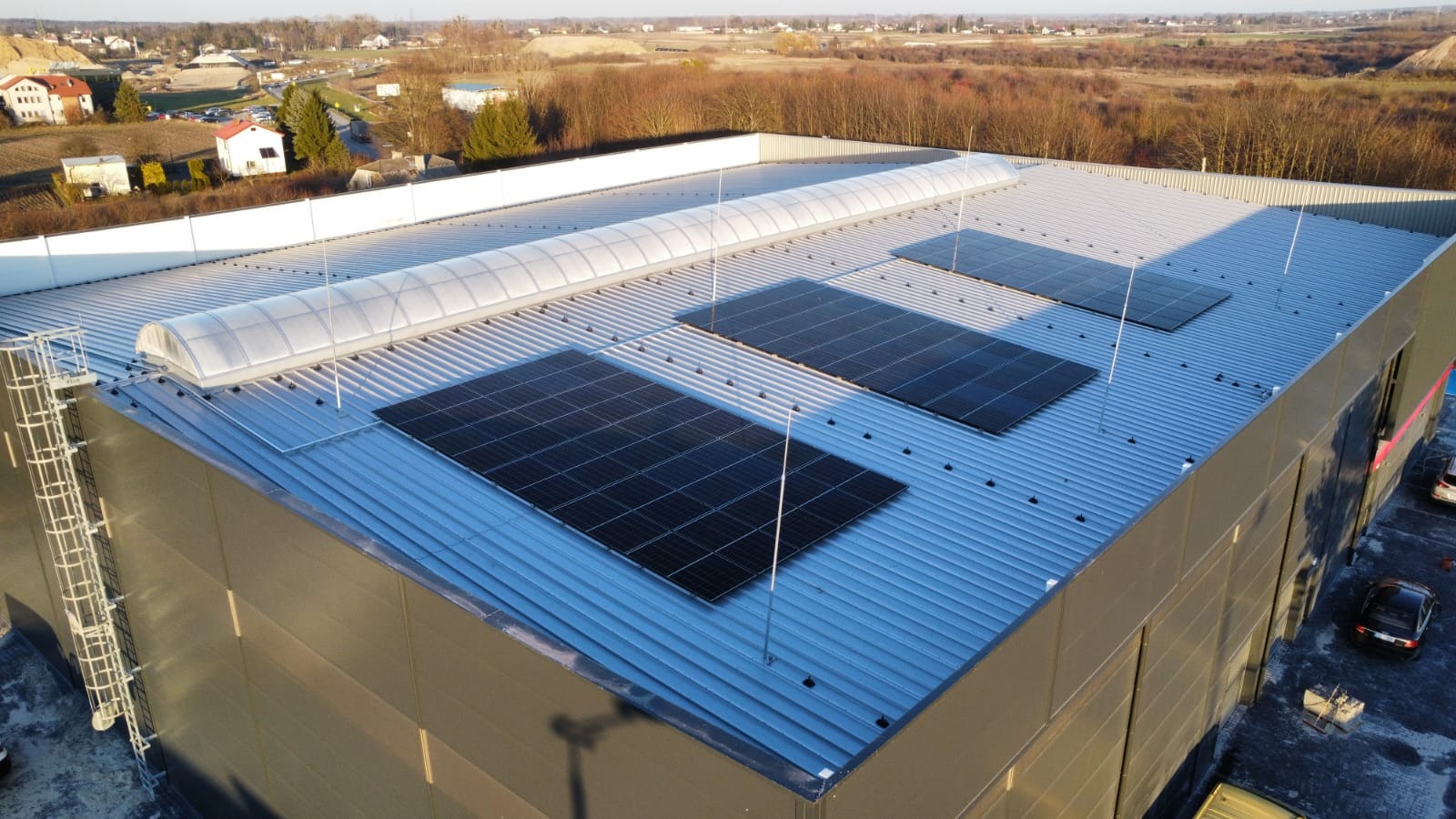 Instalacja paneli słonecznych na dachu hali przemysłowej z świetlikami. Widok z góry, w tle zabudowa mieszkalna i drzewa. Zabezpieczenia przeciw ptakom.